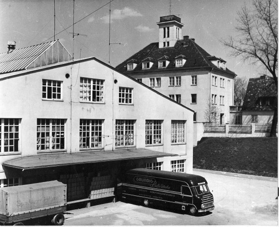 Das Schwarzweiß-Foto zeigt das ehemalige Grundigdirektionsgebäude und eine alte Fabrikhalle von außen, vor der ein schwarzer Grundig-Bus steht. 