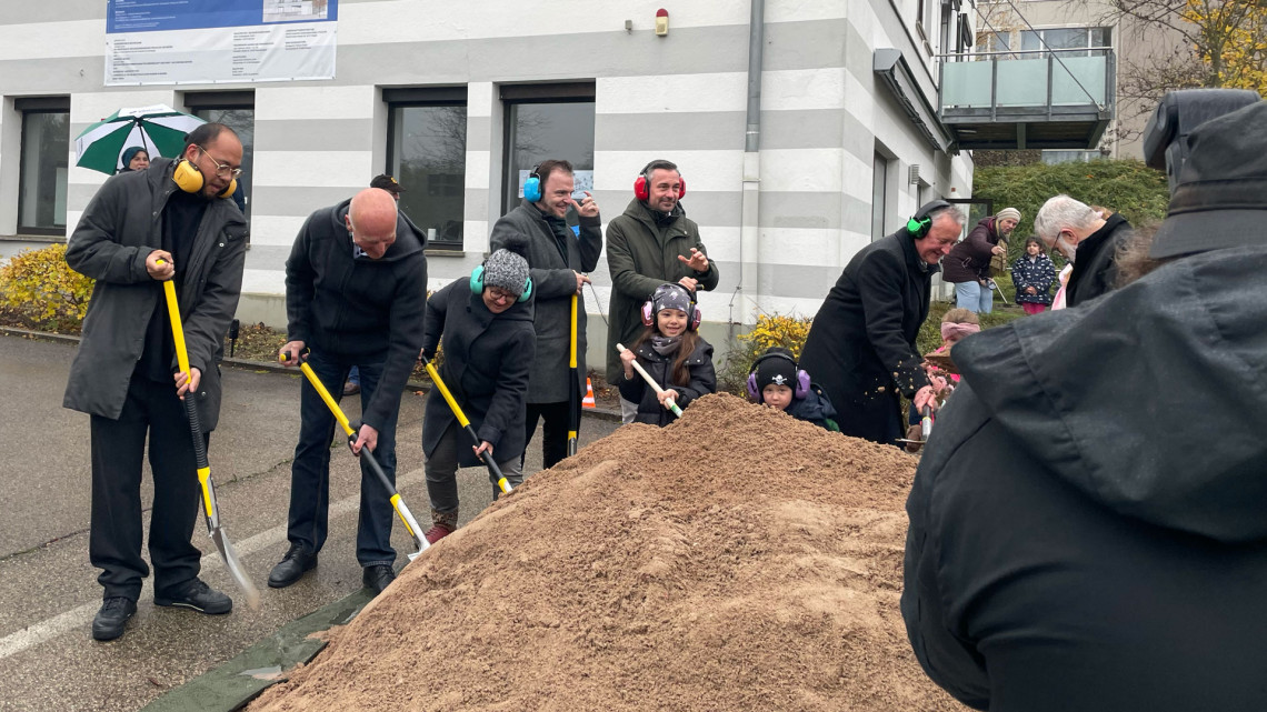 Geladene Gäste zum Spatenstich mit Schaufeln am symbolischen Sandhaufen.