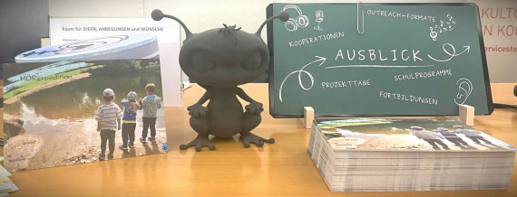 Die kleine Figur des Ausserirdischen Manuuk und eine Infotafel zur Bildungsbörse.stehen auf einem Tisch.
